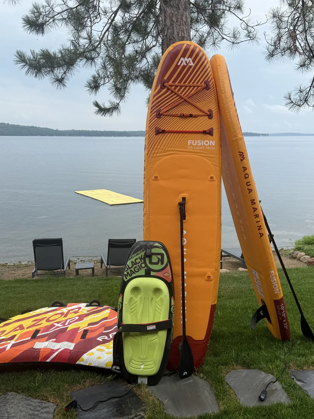 Aqua Marina Fusion paddle boards, an O'Brien kneeboard, and a towable tube on the lawn above the beach